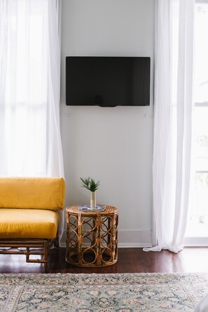 Un salón con un sofá amarillo, una mesita redonda de mimbre, un televisor de pantalla plana montado en la pared y una alfombra estampada en el suelo.