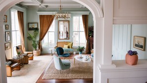 A spacious living room with a large arched doorway, a blue armchair, a sofa, a coffee table, and a chandelier. - Carr Mansion (Galveston)
