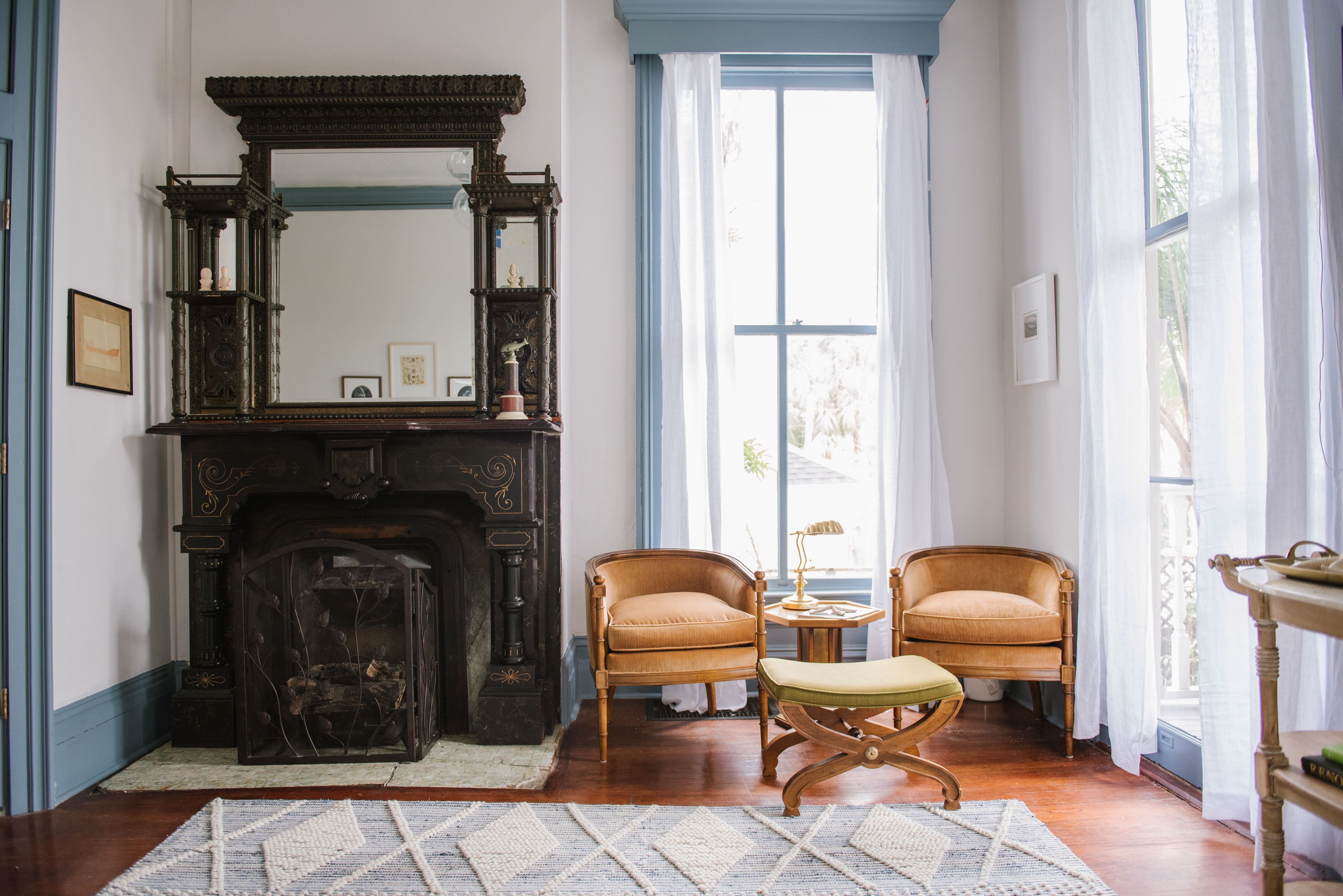 Une pièce avec une cheminée, deux fauteuils, une petite table et une fenêtre avec des rideaux.