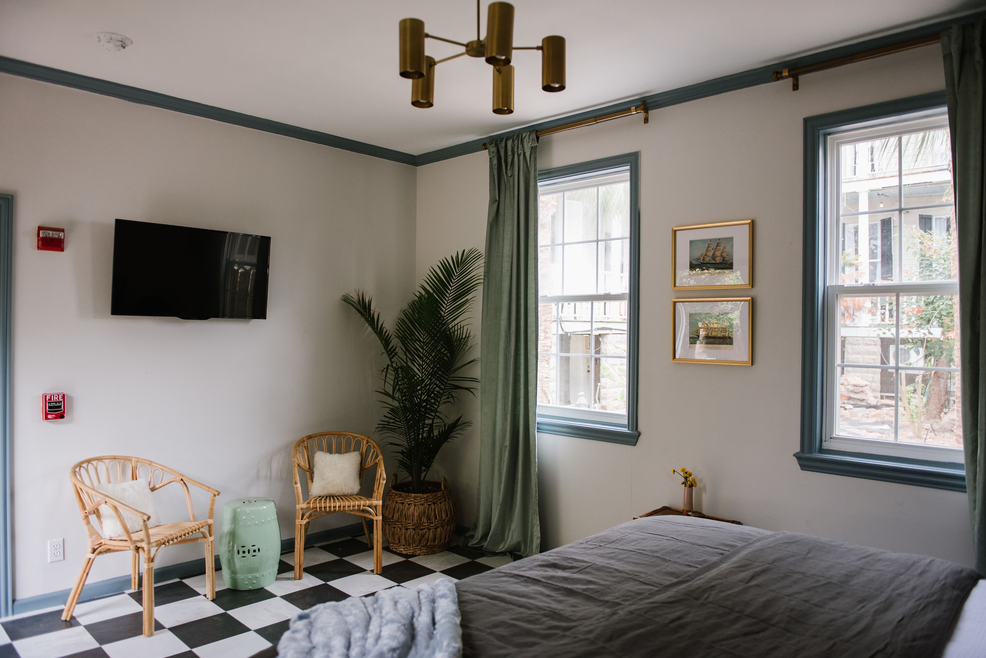 A hotel room with a checkered floor, a bed, two wicker chairs, a potted plant, a television, and framed pictures on the wall.