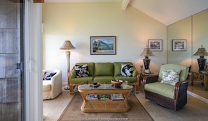 Un salon avec un canapé vert, un fauteuil en osier, une table basse en verre et deux lampes fixées au mur.