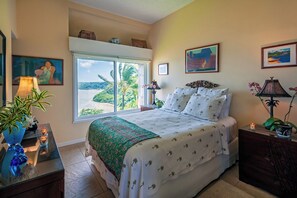 A bedroom with a large window, a bed with a green and white bedspread, a nightstand with a lamp, and a plant.