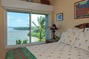 A bedroom with a large window offering a view of the ocean and palm trees. - Oceanfront and updated, top floor, perfect for honeymoon (Princeville)
