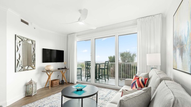 A modern living room with a large TV, a round coffee table, a sofa, and a balcony with outdoor furniture. - Beach House 413 | Oceanfront, Pet Friendly, Pools (Tybee Island)