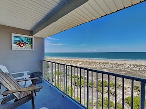 A balcony with a view of the beach, a framed painting, and outdoor furniture.