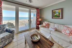 A living room with a white sofa, a patterned armchair, a wooden coffee table, and a view of the beach through large sliding glass doors.