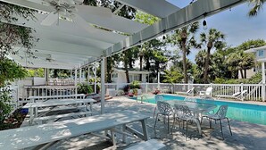 An outdoor pool area with white tables and chairs, a covered seating area, and a white fence surrounding the space. - Tybreeza A | Apartment, Shared Pool, Near Beach! (Tybee Island)