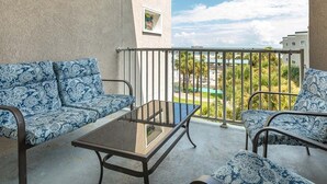 A balcony with a sofa and a table, a small window, and a view of a pool and buildings. - Tybee Beach Club 137 | Condo Overlooking Beach! (Tybee Island)