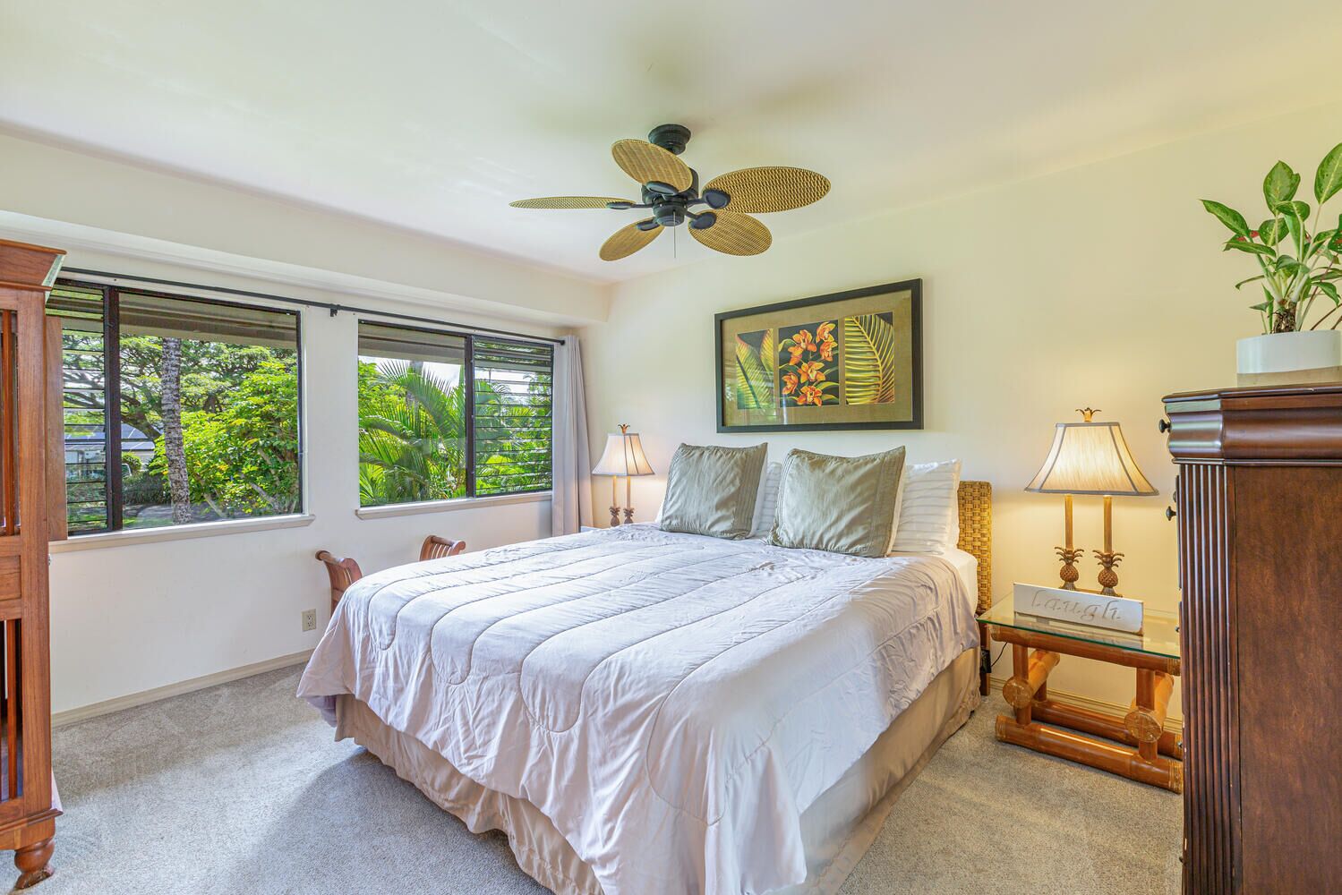 A bedroom with a large bed, two windows, a ceiling fan, a nightstand with a lamp, and a framed picture on the wall.