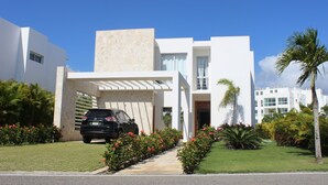 Une maison moderne à deux étages, avec une voiture garée dans l’allée, entourée de jardins bien entretenus et de palmiers.