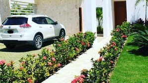 Un bâtiment moderne de couleur blanche, doté d’une allée couverte, où est garée une voiture blanche, et d’un jardin bien entretenu agrémenté de fleurs roses.