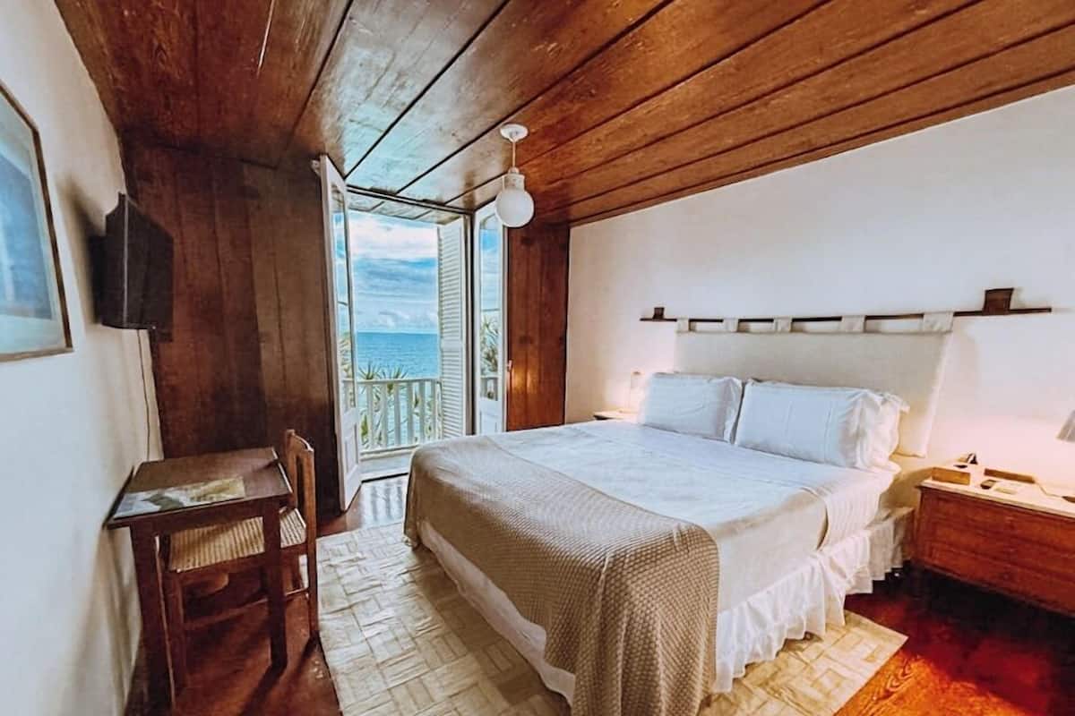 A bedroom with a bed, bedside table, chair, and a view of the ocean through an open door.