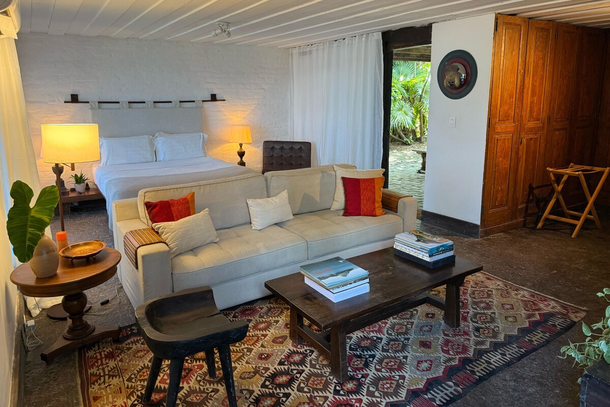A bedroom with a bed, a sofa, a coffee table, a nightstand with a lamp, a small table with a plant, and a view of the outside.