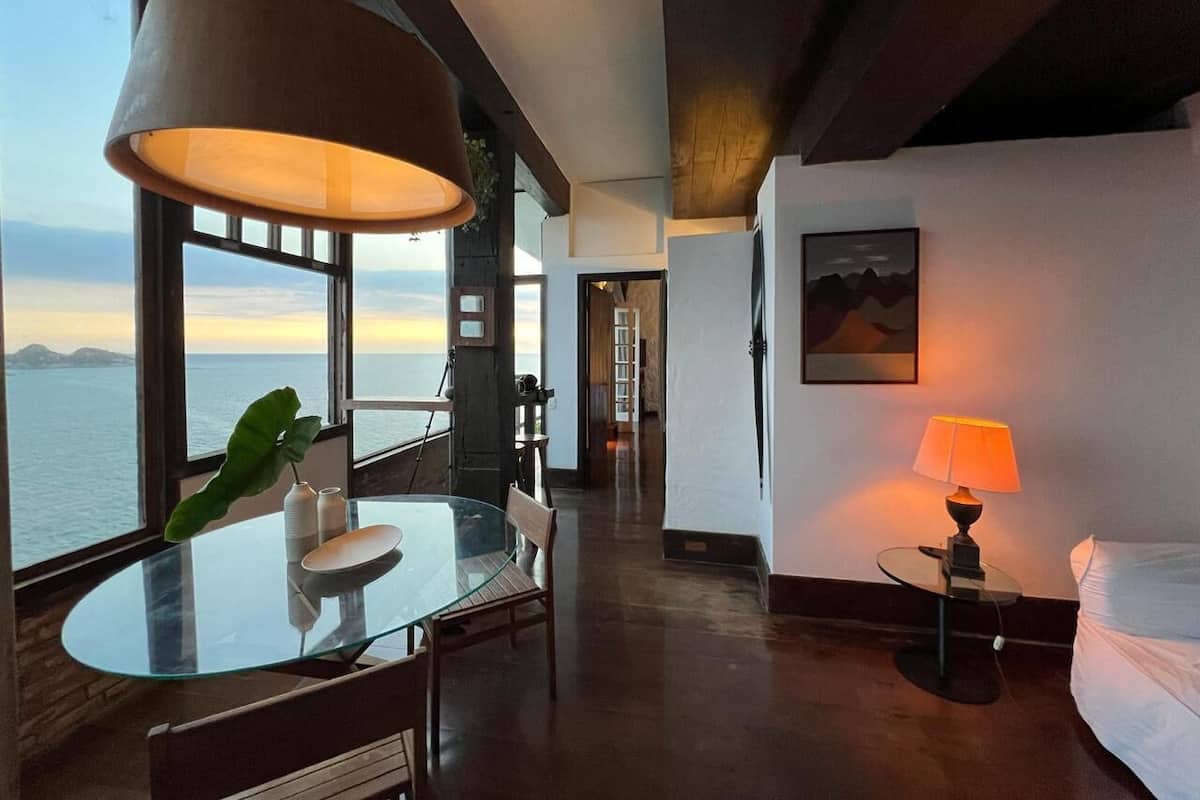 A modern hotel room with a glass table, a large window with a view of the sea, a bed, and a lamp.