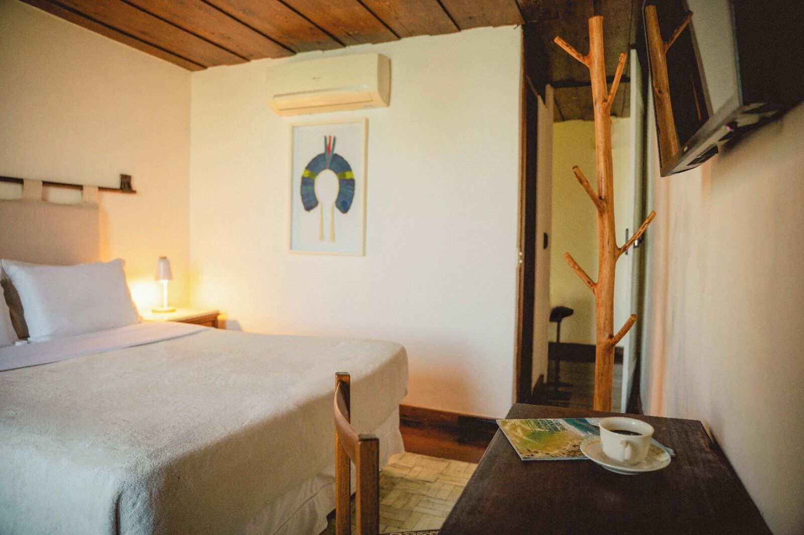 Quarto com cama, mesa de cabeceira com xícara e pires, ar condicionado fixado na parede e um quadro emoldurado.