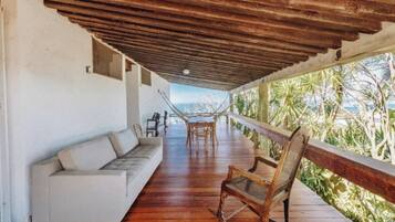A covered porch with a sofa, rocking chair, and dining table, overlooking a tropical landscape.