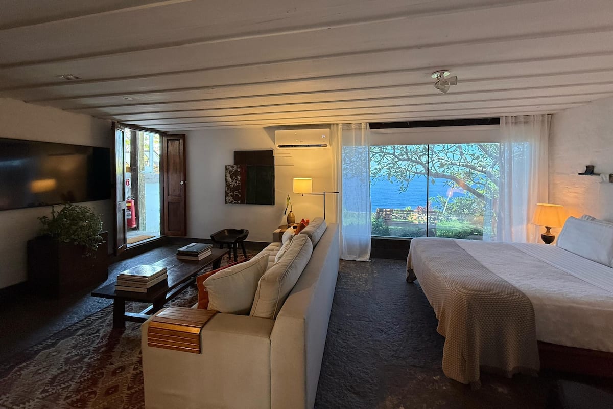 A room with a bed, a sofa, a TV, a table with books, a plant, and a view of the outdoors.