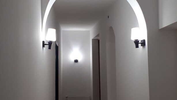 A hallway with white walls, a wooden floor, and two wall-mounted light fixtures on either side. - Gnt Plus (Tbilisi)