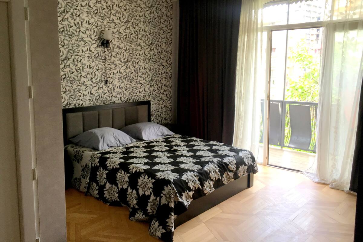 A bedroom with a bed, a patterned wall, a window with curtains, and a wooden floor.