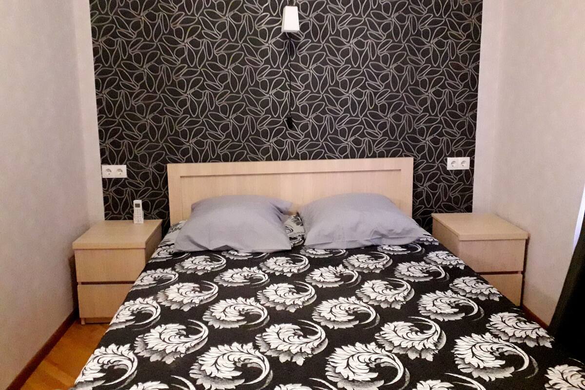 A small, neatly made bed with a patterned bedspread, two bedside tables, and a wall with a leaf pattern.