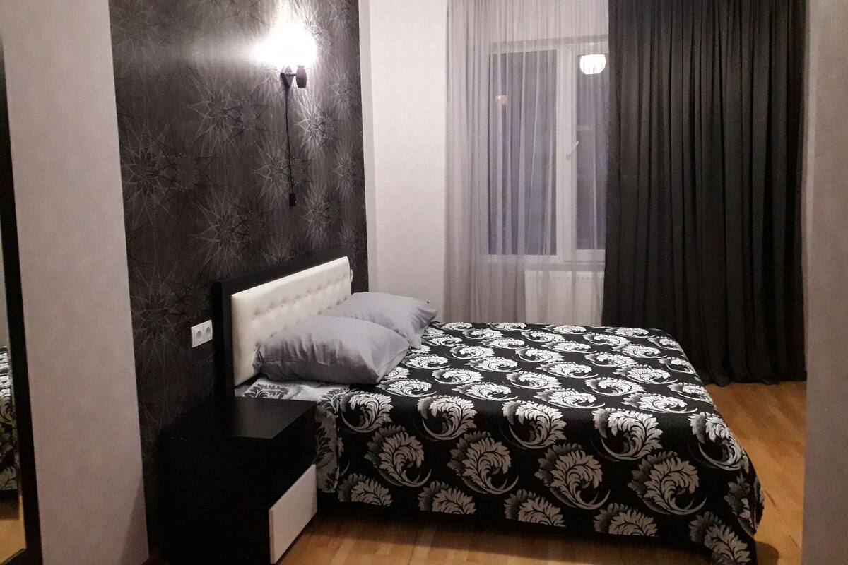A bedroom with a bed, a nightstand, a wall-mounted light, a window with sheer curtains, and a dark curtain.