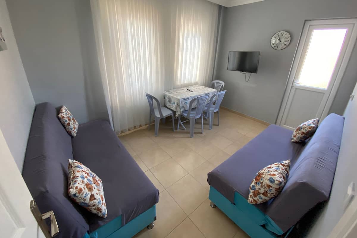 A room with two blue sofas with floral cushions, a small table with chairs, a TV on the wall, and a door with a window.