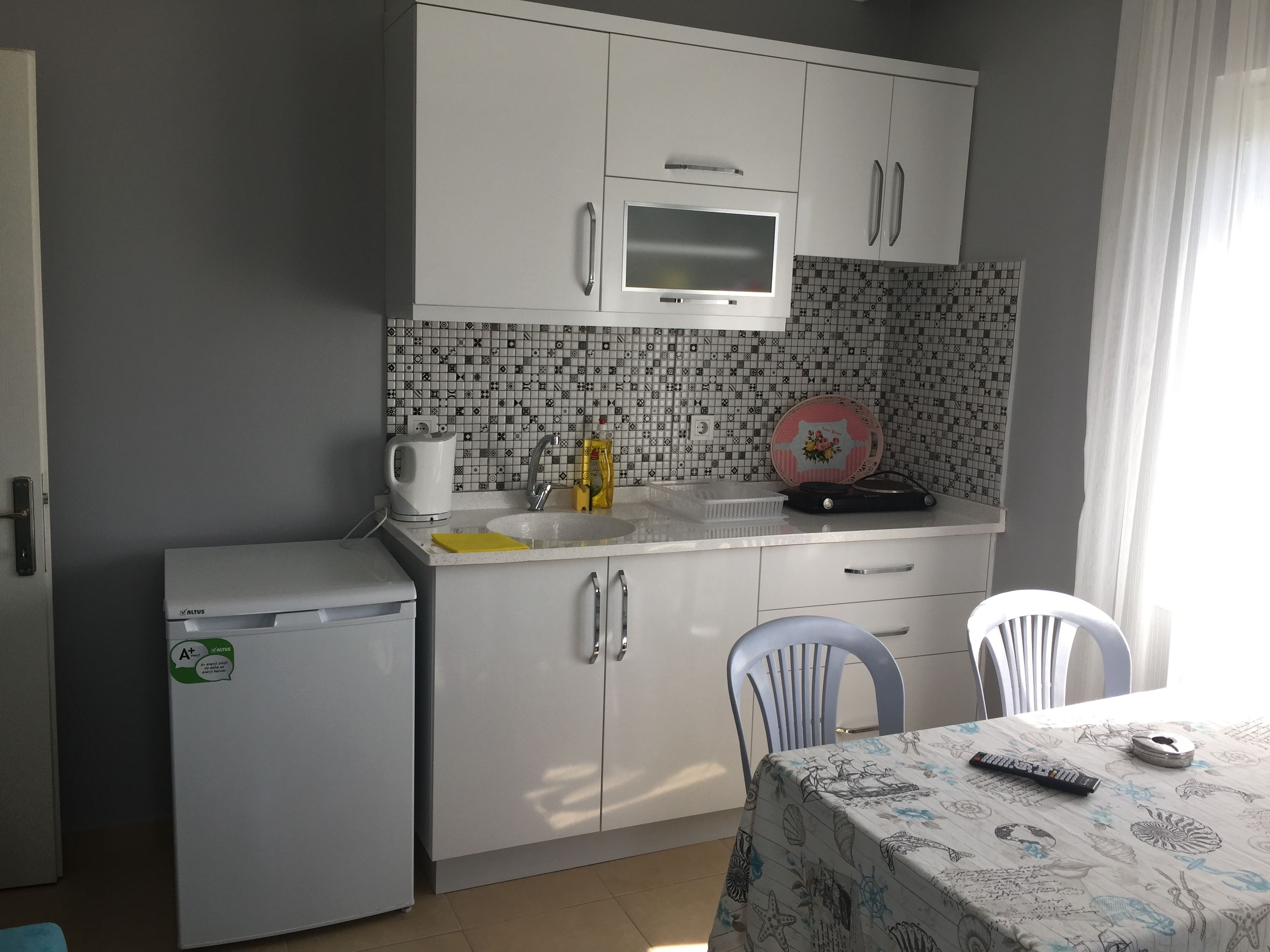 A compact kitchen with a refrigerator, microwave, and sink. There is a table with a tablecloth and a remote control on it.
