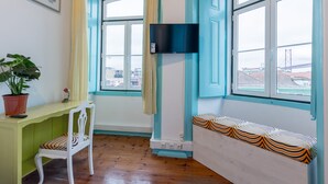 Une pièce avec un plancher en bois, une chaise blanche, un bureau vert, une plante en pot, un téléviseur fixé au mur et un banc avec un coussin à rayures.