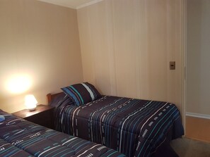 A hotel room with two beds, a nightstand, a lamp, and a door with an intercom. - Departamento Edificio Parque Pacifico (Concon)