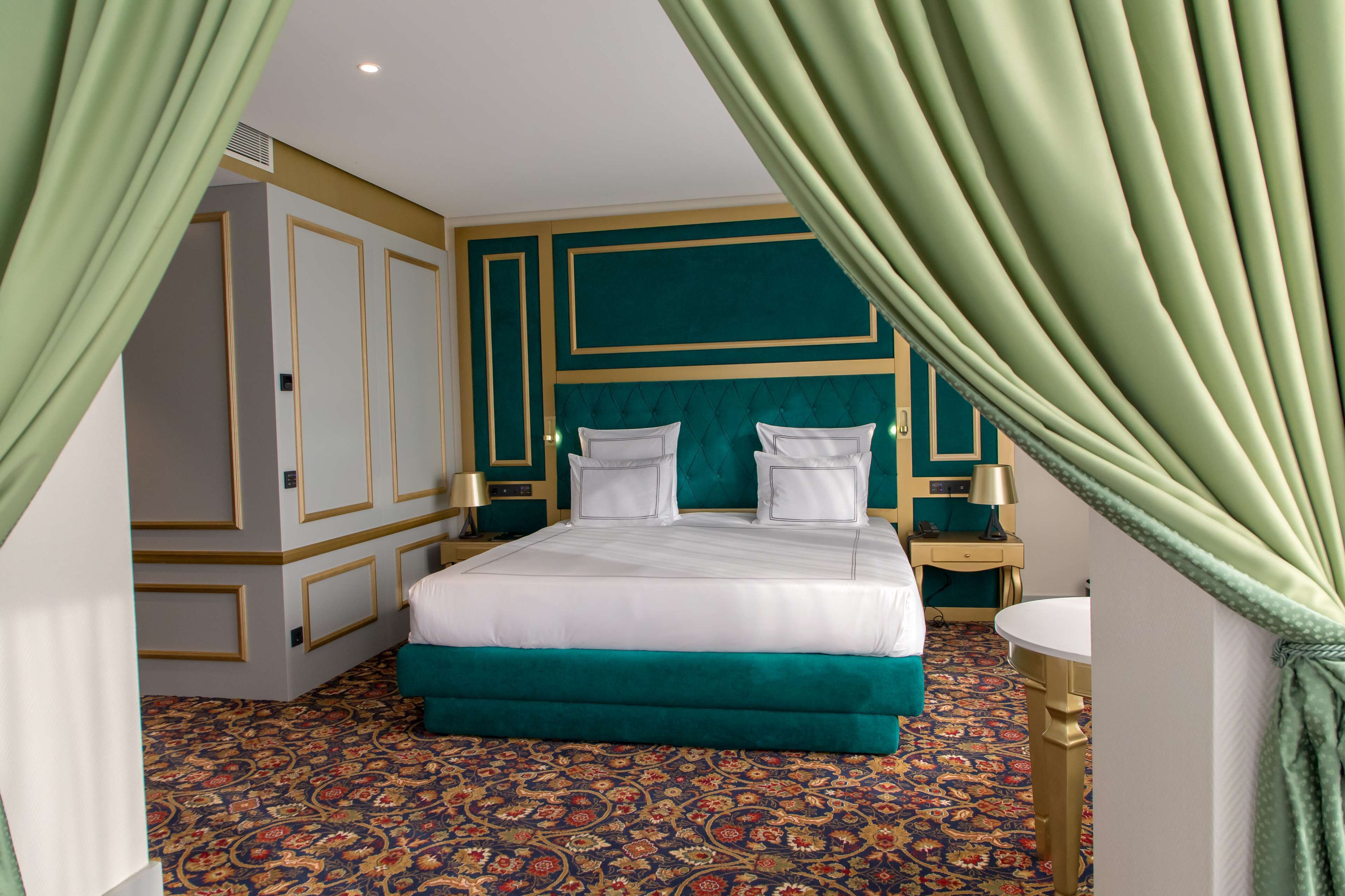 A hotel room with a large bed, green curtains, a patterned carpet, and a small bedside table with a lamp.