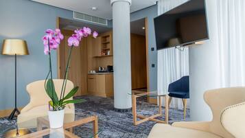 Habitación de hotel moderna con una zona de estar que incluye una mesa de centro de vidrio, un sofá color beige y un sillón azul. Se observa un televisor de pantalla plana montado en la pared, una orquídea en maceta y un aparador de madera al fondo.