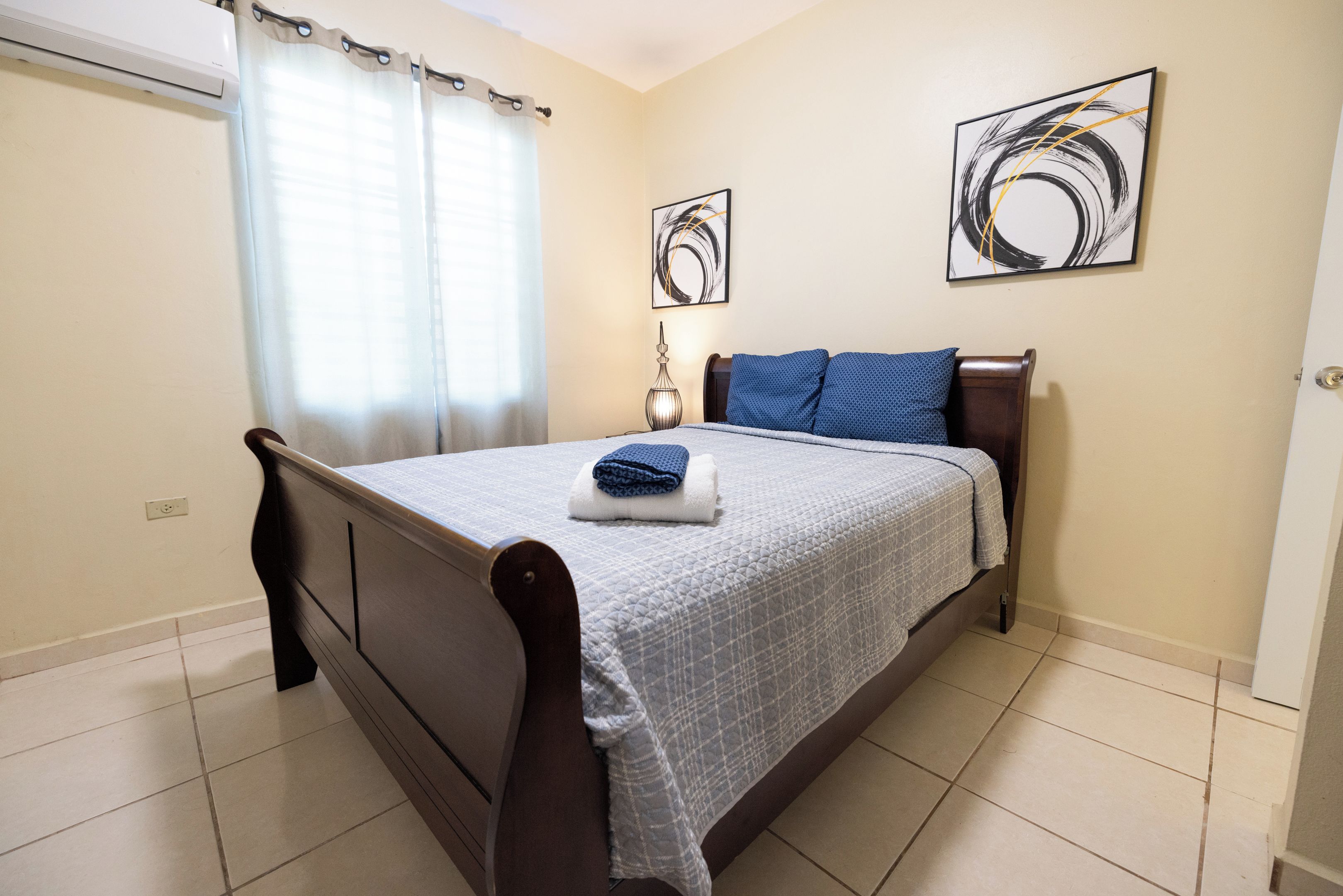 Un dormitorio con cama, cortinas, una mesita de noche y dos cuadros en la pared.