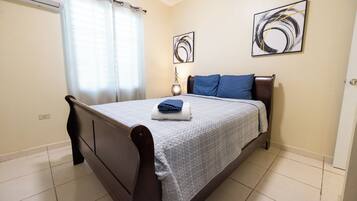 Un dormitorio con cama, cortinas, una mesita de noche y dos cuadros en la pared.