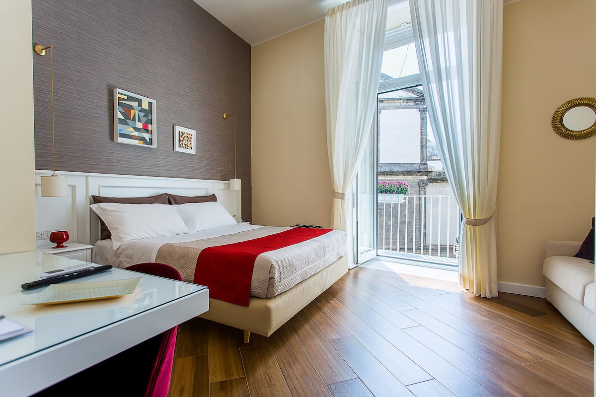 A modern hotel room with a bed, a glass desk, a red throw blanket, and a view of the outside.