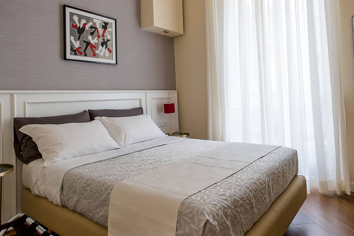A hotel room with a bed, a headboard, a bedside table, a nightstand, a wall-mounted lamp, a window with curtains, and a framed picture on the wall.