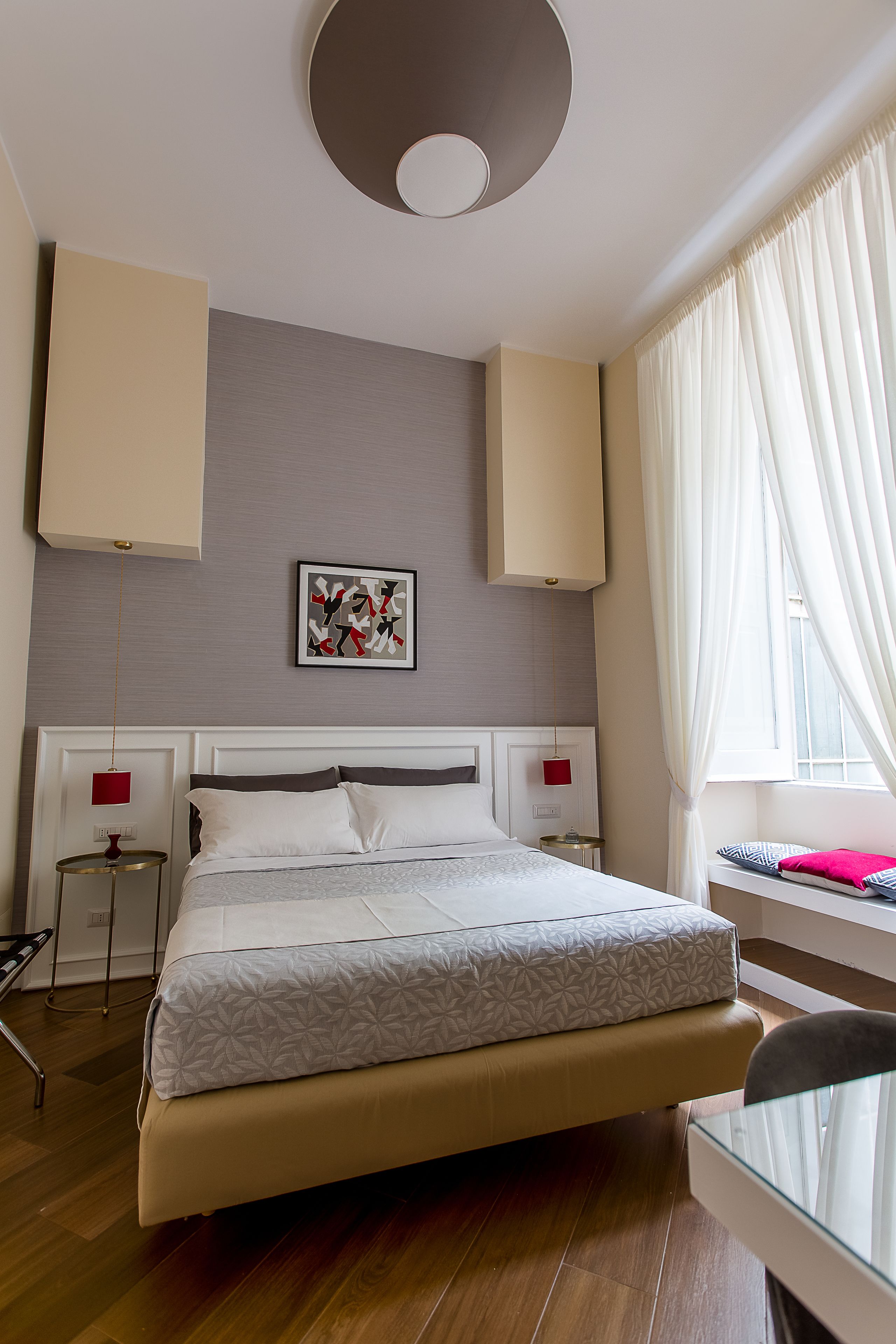 A modern hotel room with a large bed, a bedside table, a chair, a window with curtains, and a wall-mounted artwork.