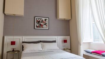 A modern hotel room with a large bed, a bedside table, a chair, a window with curtains, and a wall-mounted artwork.