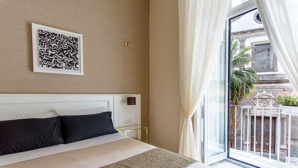 A hotel room with a large bed, a framed artwork on the wall, and a view of the city through a balcony. - NovaNova (Naples)
