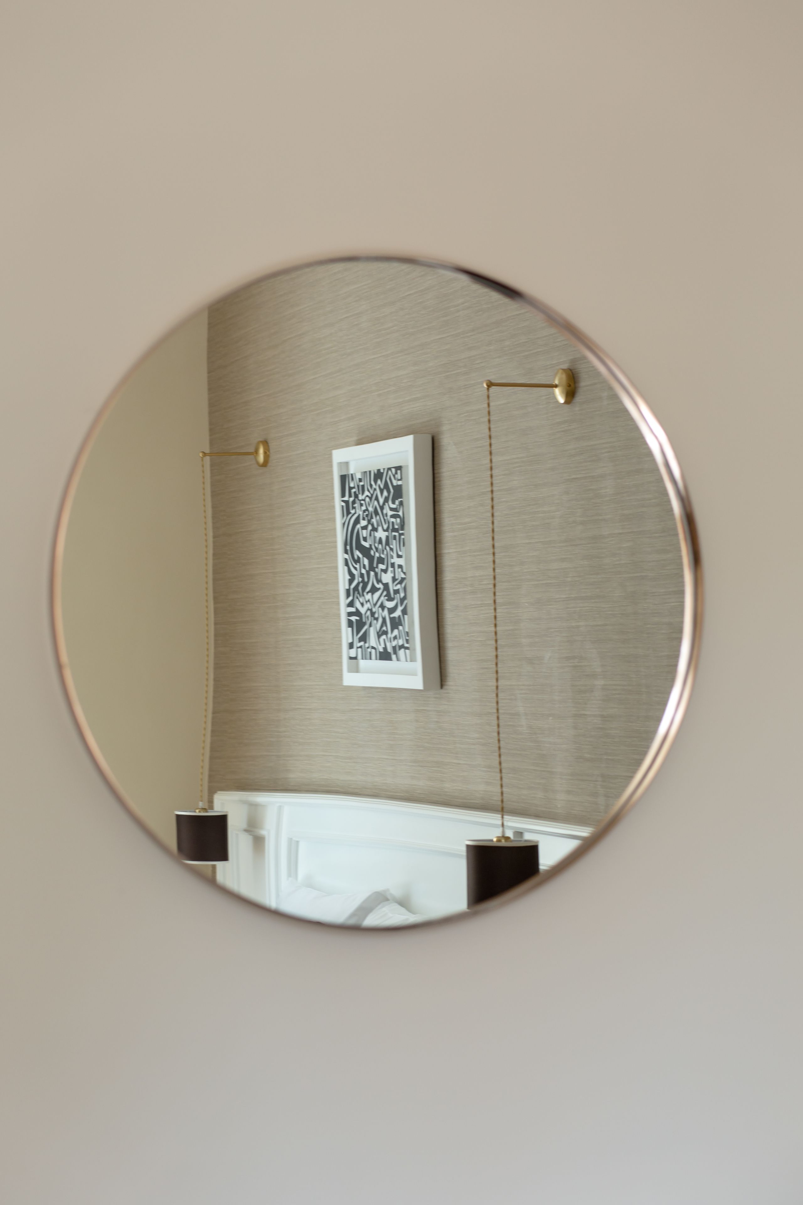 Un espejo circular que refleja una habitación con un cuadro de motivos decorativos en la pared, una lámpara de pie negra y un mueble blanco.