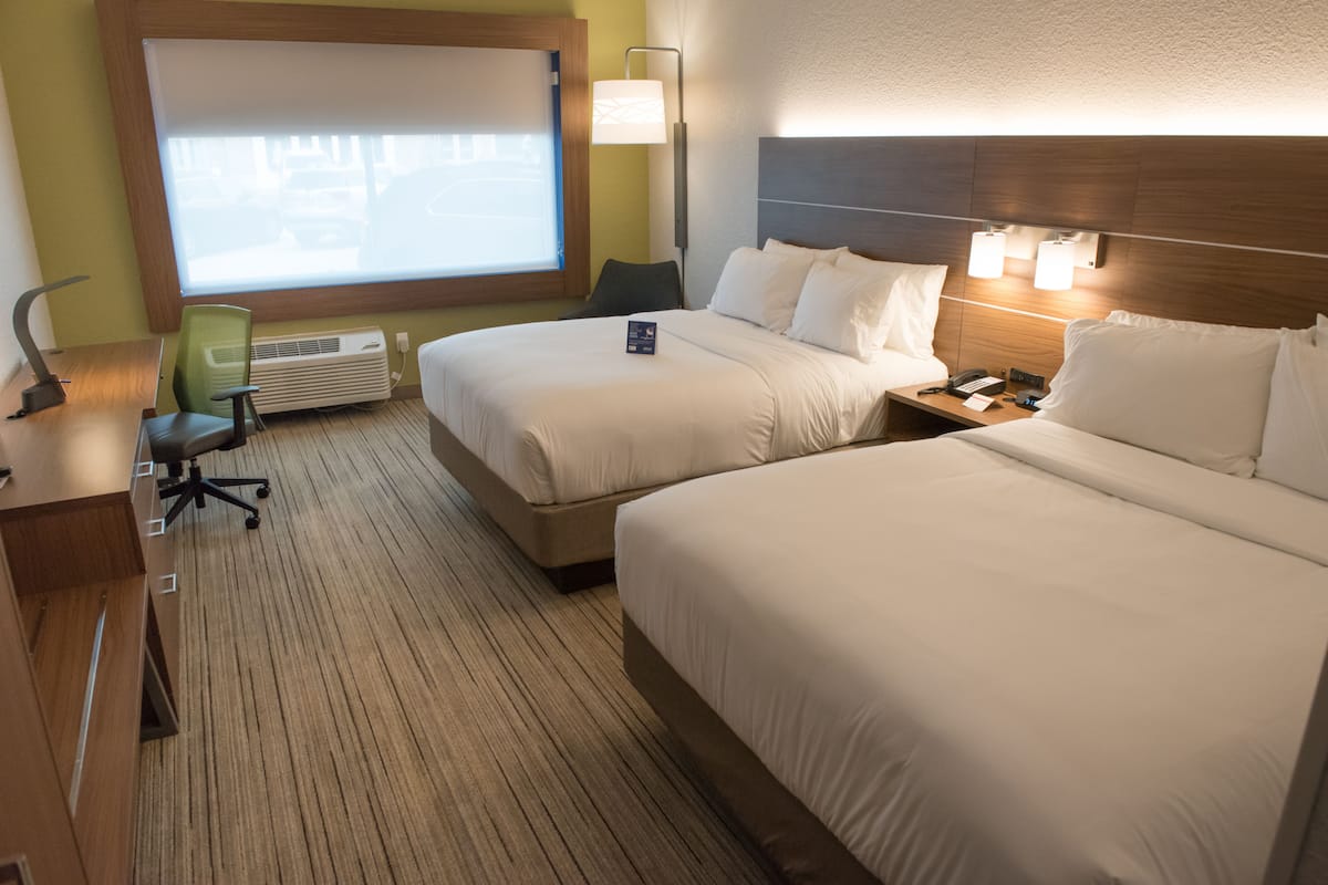 A hotel room with two beds, a desk, a chair, a lamp, and a window with blinds.