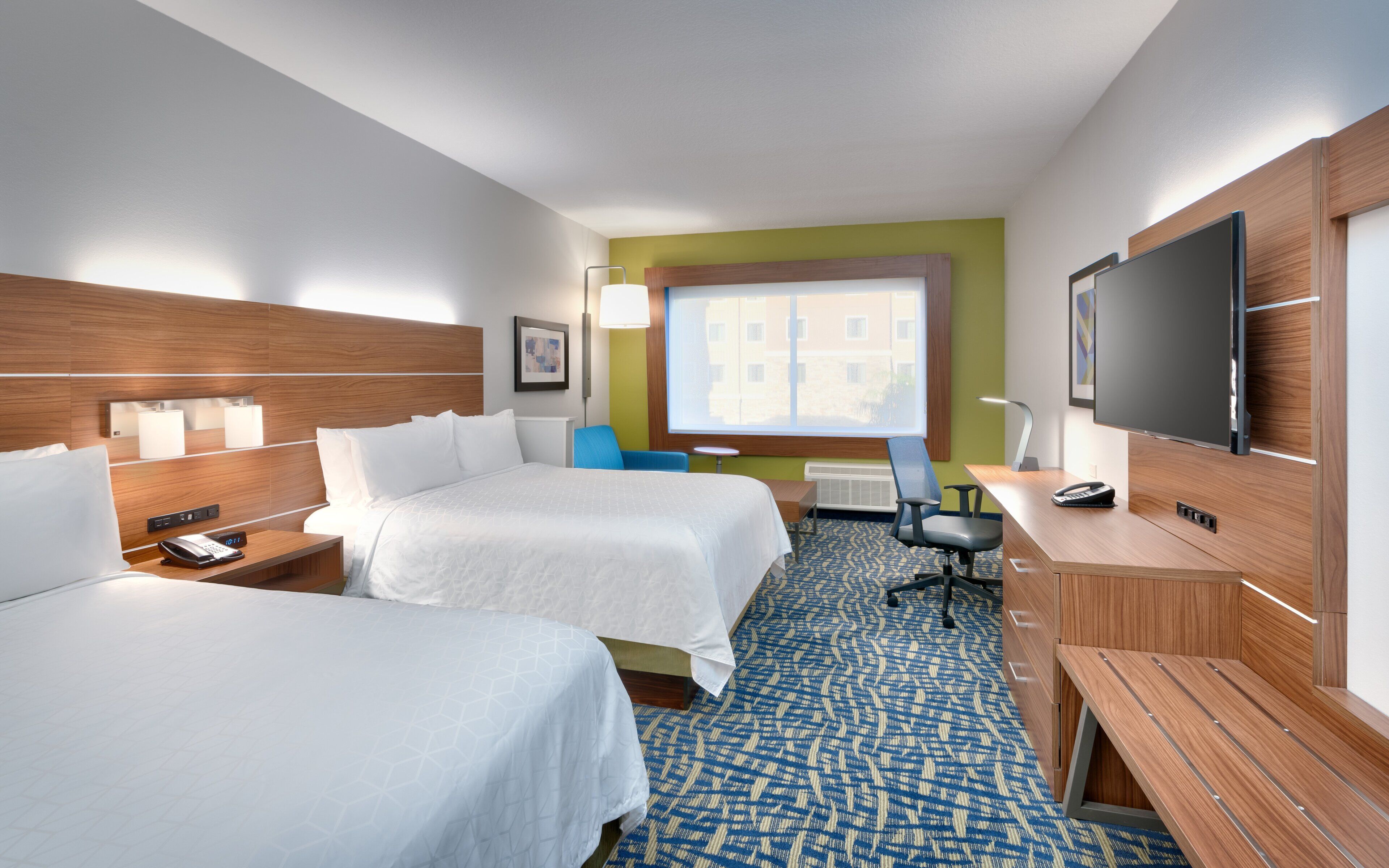 A hotel room with two beds, a desk, a chair, a television, and a window with a view of buildings.