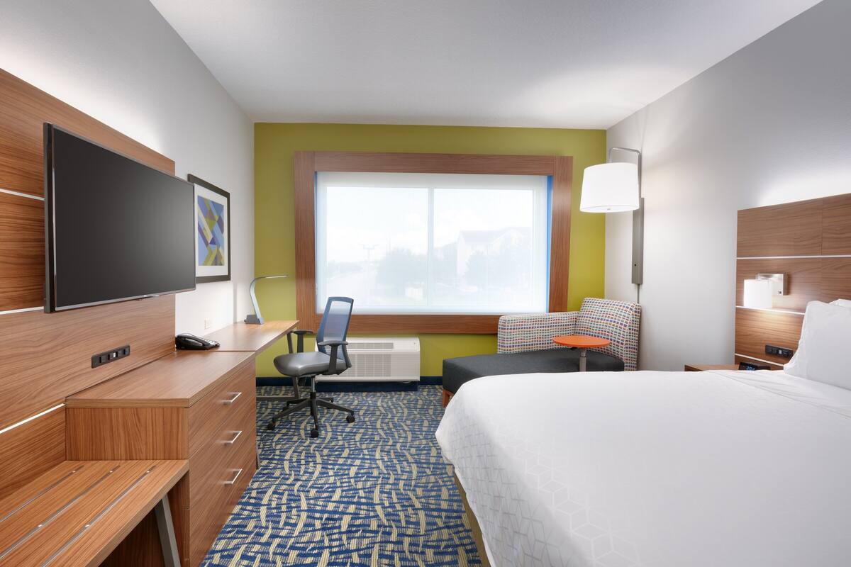 A hotel room with a bed, a desk with a chair, a television, and a window with a view of buildings.