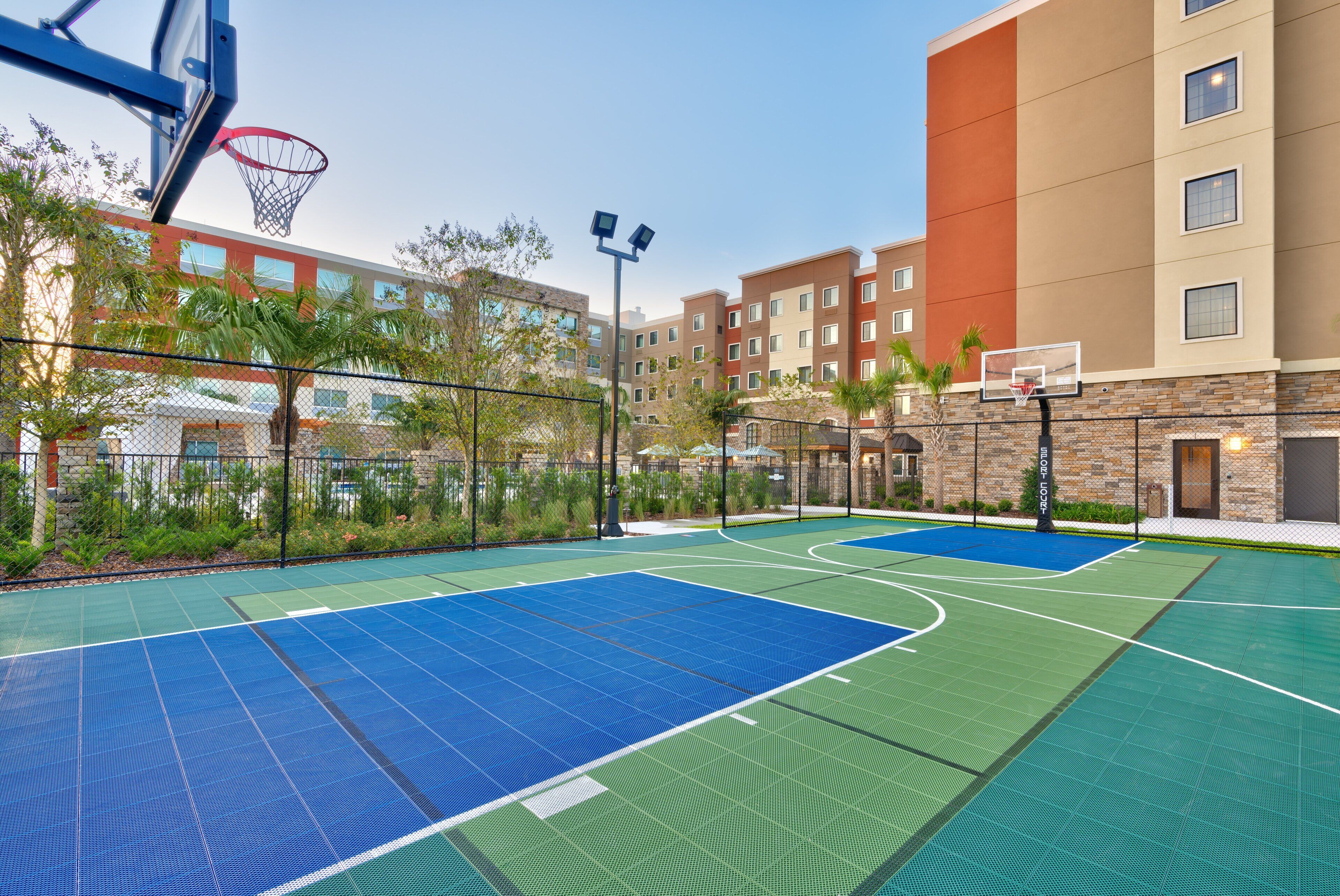 An outdoor basketball court with two hoops, surrounded by a chain-link fence and palm trees.