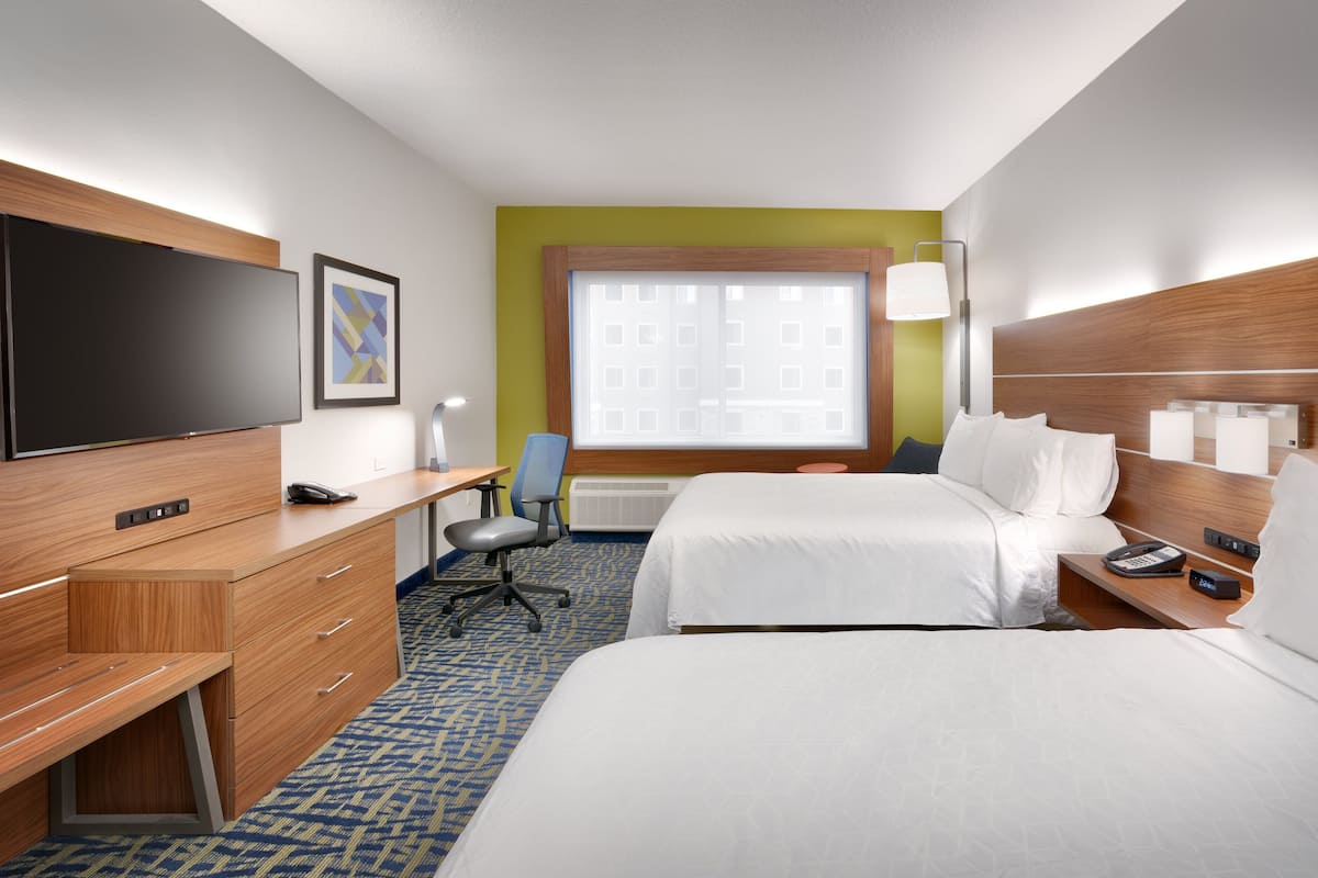 A hotel room with a bed, desk, chair, TV, and a window with a view of buildings.