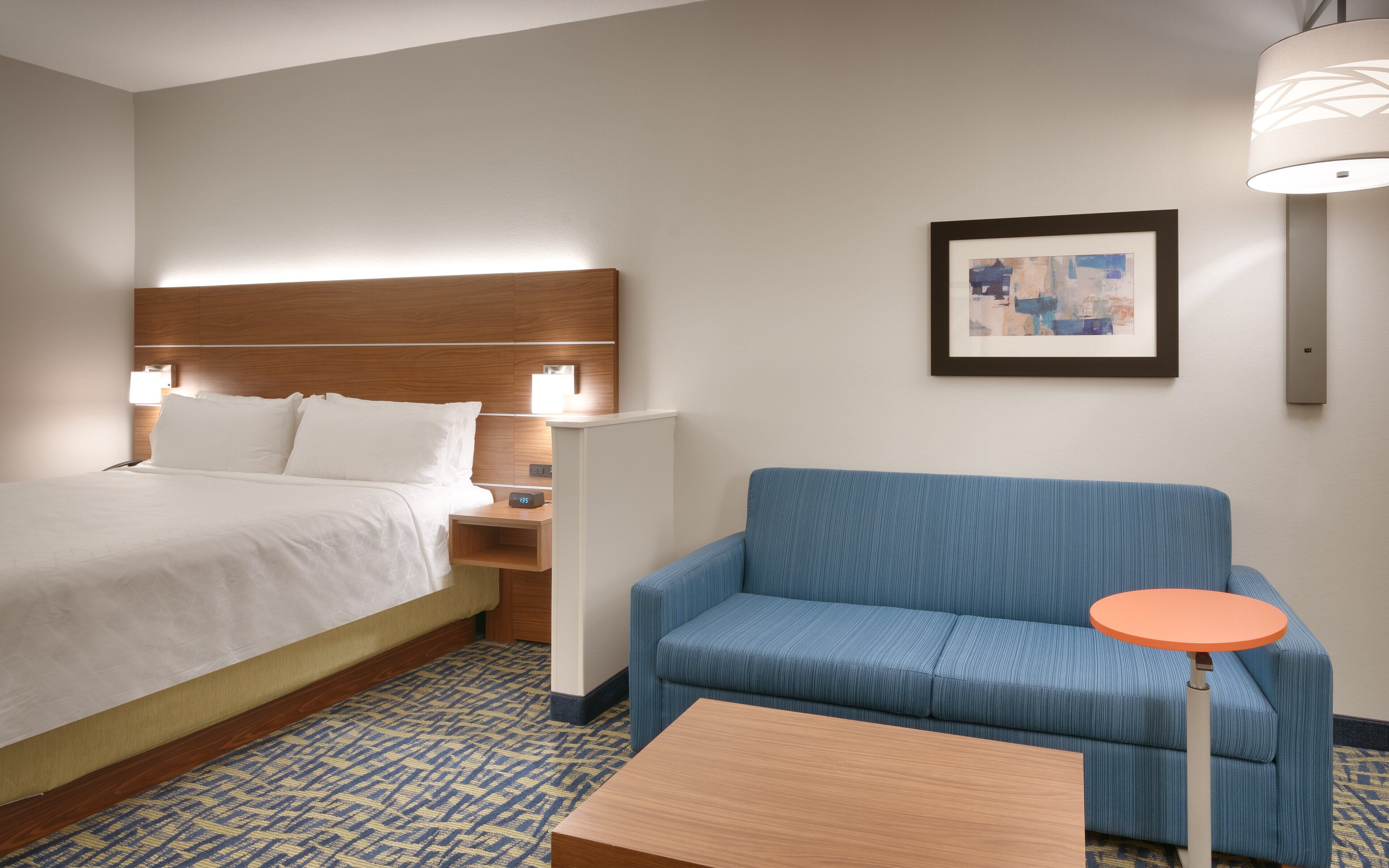 A hotel room with a bed, a nightstand, a blue sofa, a small table, and a framed picture on the wall.