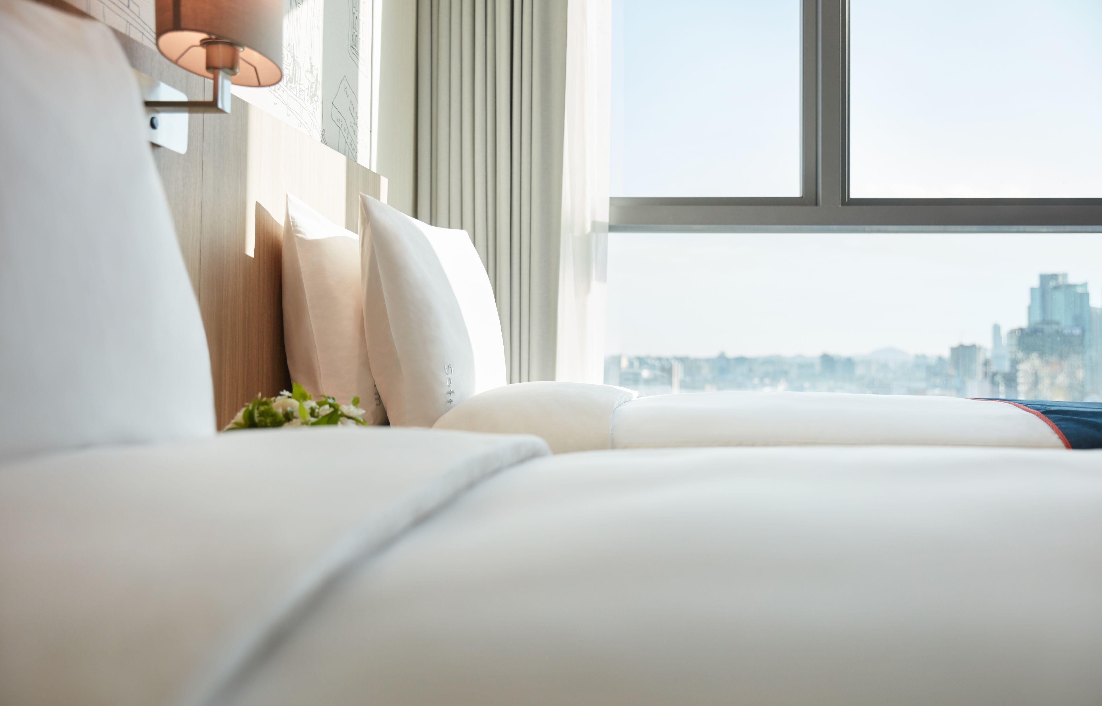 A hotel room with a large window, a bed with white bedding, and a lamp on the wall.