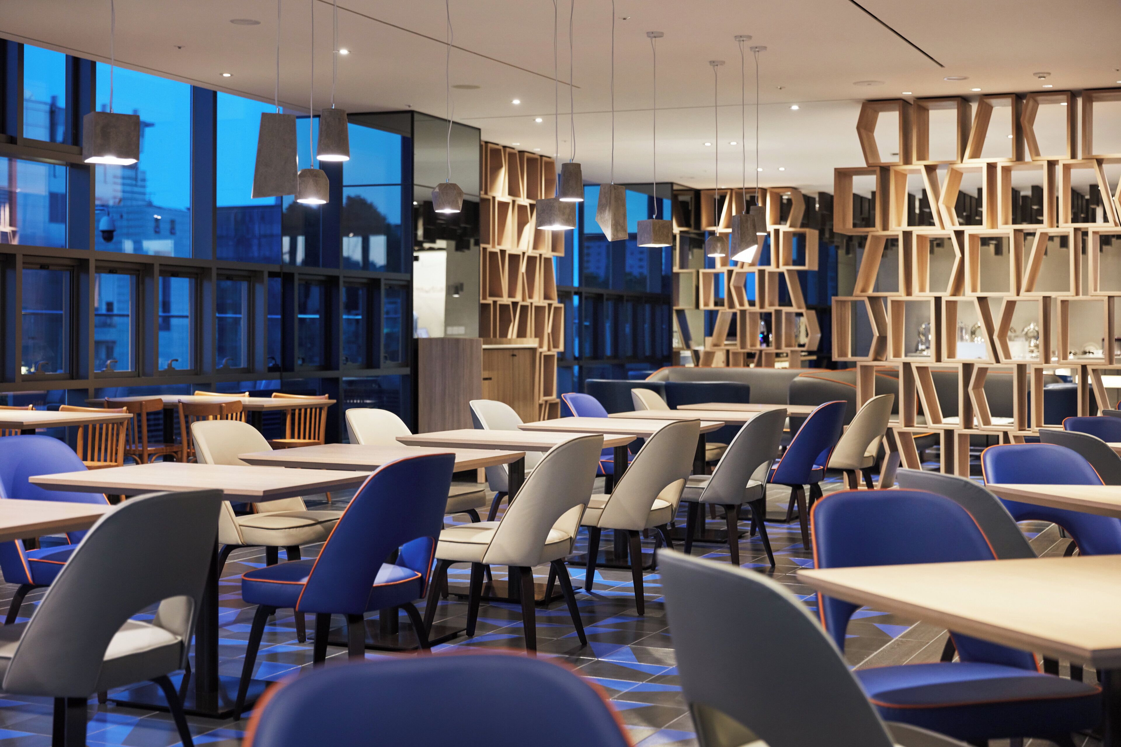 A modern dining area with tables and chairs, large windows, and a geometric bookshelf structure.