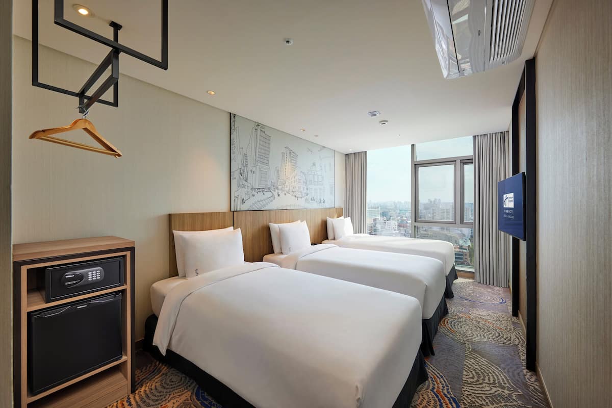 A hotel room with three beds, a minibar, a TV, and a city view.