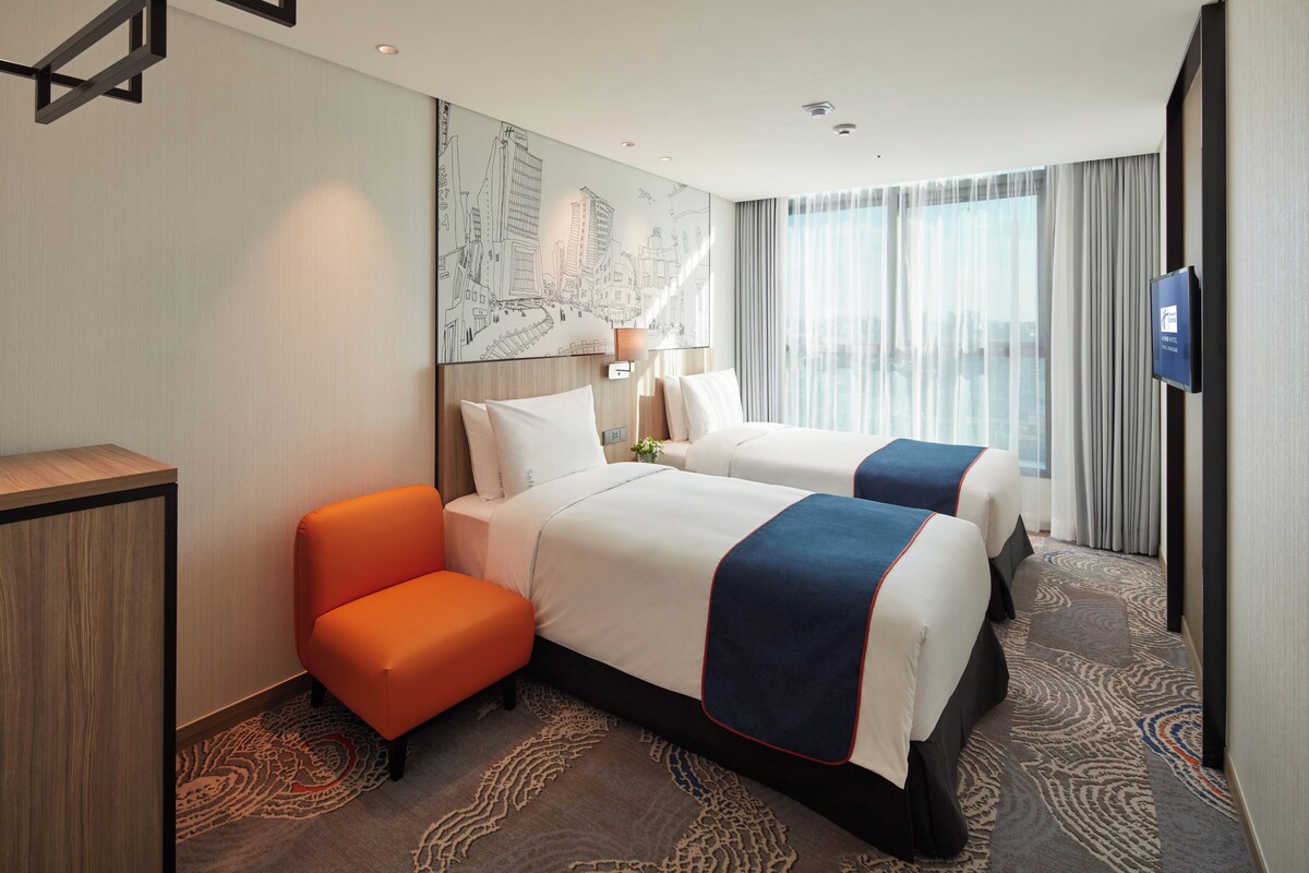 Een hotelkamer met twee bedden, een bureau, een oranje stoel, een televisie en een groot raam met gordijnen.