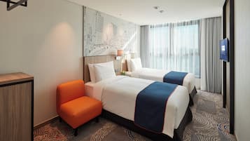 Une chambre d’hôtel avec deux lits, un bureau, une chaise orange, une télévision et une grande fenêtre avec des rideaux.